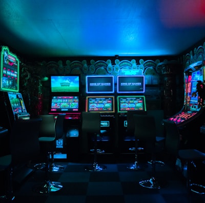 Dimly lit gaming arcade featuring a variety of brightly illuminated slot machines and video gaming screens. Neon signs and colorful displays create a vibrant atmosphere. The arrangement of gaming machines suggests an inviting space for entertainment, with chairs positioned for players.
