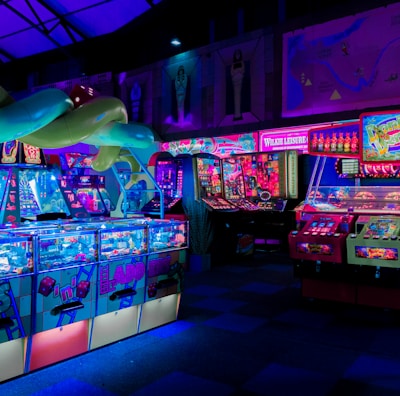 An indoor arcade filled with vibrant gaming machines lit with neon blue and pink lights. The scene includes various arcade games with flashing screens and colorful displays, and a large overhead structure resembling a green snake. The background shows illuminated signs and themed decor, contributing to an immersive entertainment atmosphere.