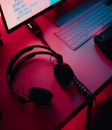 A dimly lit gaming setup with a monitor displaying a game's menu on the screen. A mechanical keyboard sits in front of the monitor. A pair of over-ear headphones and a game controller rest on the desk. The scene is bathed in a red and blue light, creating a moody gaming atmosphere.