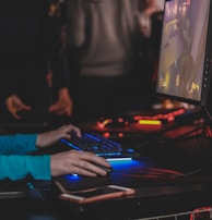 A person is engaged in a video game at a computer setup, using a keyboard and mouse. The scene is dimly lit, with the glow from the monitor reflecting off the hands of the player. Several other people are present in the background, indicating a social or competitive gaming environment.