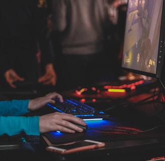 A person is engaged in a video game at a computer setup, using a keyboard and mouse. The scene is dimly lit, with the glow from the monitor reflecting off the hands of the player. Several other people are present in the background, indicating a social or competitive gaming environment.