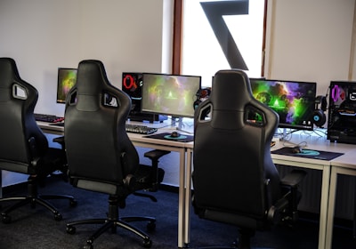A row of gaming stations equipped with large monitors, gaming chairs, keyboards, and headphones. The setup is organized and ready for gaming, featuring a colorful screen display. The room has a modern and tech-savvy ambiance.