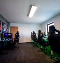 A well-lit gaming room features multiple gaming stations with high-backed chairs and monitors. The room has a minimalist setup with green and black color tones. Large posters adorn the walls, and the room appears clean and organized.