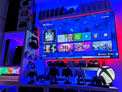 A modern gaming setup featuring a large wall-mounted screen displaying a gaming console interface. The room is decorated with gaming-themed items such as controllers and collectibles. The ambient lighting includes shades of blue and red, creating a vibrant atmosphere.