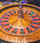A roulette wheel with alternating red and black numbered pockets on a wooden platform. The wheel contains a small white ball resting in one of the pockets, indicating gameplay. Surrounding the wheel are stacks of colorful poker chips, suggesting a casino environment.