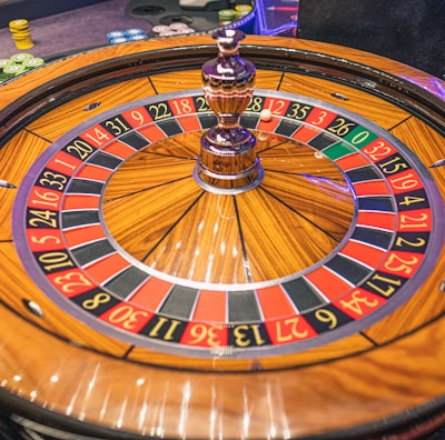 A roulette wheel with alternating red and black numbered pockets on a wooden platform. The wheel contains a small white ball resting in one of the pockets, indicating gameplay. Surrounding the wheel are stacks of colorful poker chips, suggesting a casino environment.