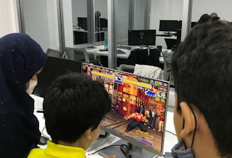 A group of children is gathered around a desktop computer, engaged in playing a colorful, action-packed arcade game displayed on the monitor. The setting appears to be a modern, well-lit office or gaming space with large glass windows, showing multiple computer workstations in the background.
