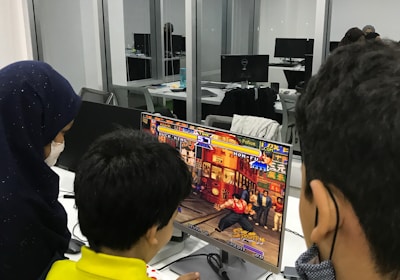 A group of children is gathered around a desktop computer, engaged in playing a colorful, action-packed arcade game displayed on the monitor. The setting appears to be a modern, well-lit office or gaming space with large glass windows, showing multiple computer workstations in the background.