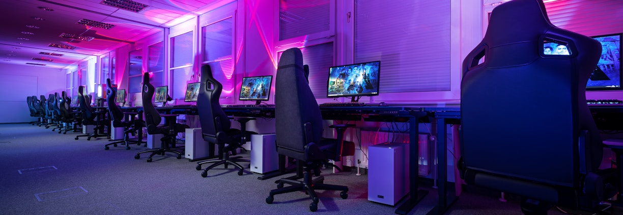 A row of gaming computers with large monitors and racing-style chairs is set up along the interior of a modern room. The space is illuminated with vibrant purple and blue LED lighting, creating a futuristic atmosphere. The room is organized and clean, suggesting a professional gaming or esports environment.