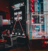 An arcade setting featuring a classic Space Invaders game setup and a modern gaming machine with seats. Bright neon lights illuminate the area, with scores displayed on digital screens. A bright and visually engaging environment full of electronic components.