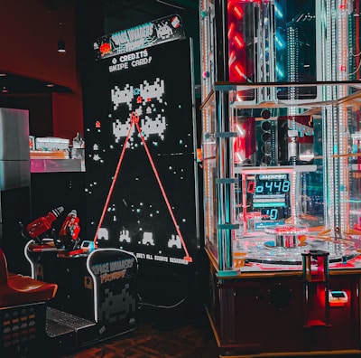 An arcade setting featuring a classic Space Invaders game setup and a modern gaming machine with seats. Bright neon lights illuminate the area, with scores displayed on digital screens. A bright and visually engaging environment full of electronic components.