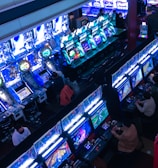 A bustling arcade filled with rows of gaming machines, each with vibrant displays and controls. Several people are seated at these machines, deeply engaged in playing games. Bright fluorescent lights cast a blue and neon glow across the room, enhancing the lively and energetic atmosphere.