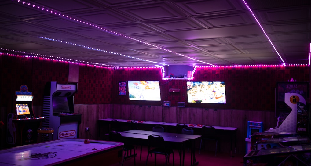 A dimly lit room with vibrant purple LED strip lights on the ceiling, featuring multiple gaming stations. There are two large screens showing video games, along with various arcade machines, including a Nintendo arcade cabinet. An air hockey table is positioned in the foreground.
