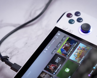A handheld gaming console with a white body and colorful buttons features a screen displaying various game icons. The device is connected to a power source via a black cable and rests on a marble surface. The interface shows games like Apex and Street Fighter.