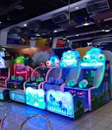 A brightly lit arcade filled with colorful gaming machines sits under ceiling lights. Neon lights illuminate various game stations, creating a playful and vibrant environment. A few red chairs are scattered around the reflective, tiled floor, and a person is observing one of the machines. The atmosphere is lively and cheerful, featuring games with cartoonish designs and engaging themes.