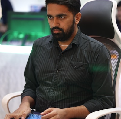 A person is seated in a chair, focused intently on something, with hands positioned on a gaming console or arcade controller. The setup appears to be indoors, with visible screens and lighting in the background, suggesting an electronic gaming environment.